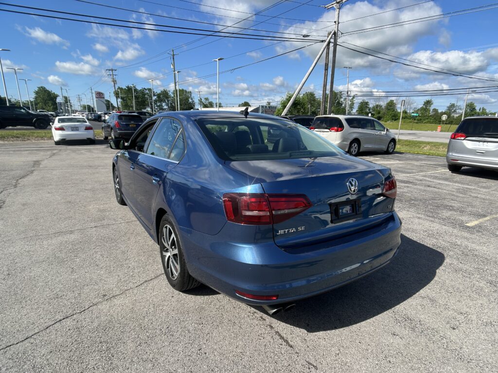 Blue Volkswagen Jetta SE in a parking lot, with other cars and tall power lines overhead under a blue sky with clouds.