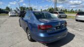 Blue Volkswagen Jetta SE in a parking lot, with other cars and tall power lines overhead under a blue sky with clouds.