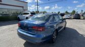 Blue sedan in the foreground at a car dealership lot with other cars and a white building on the left.
