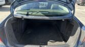 Open blue car trunk showing a spacious, dark carpeting cargo area with rear seats visible and the trunk lid raised; shows Volkswagen logo on floor mat.