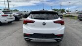 Rear view of a white Buick Encore GX parked in a lot among other cars, with power lines overhead and a blue sky.