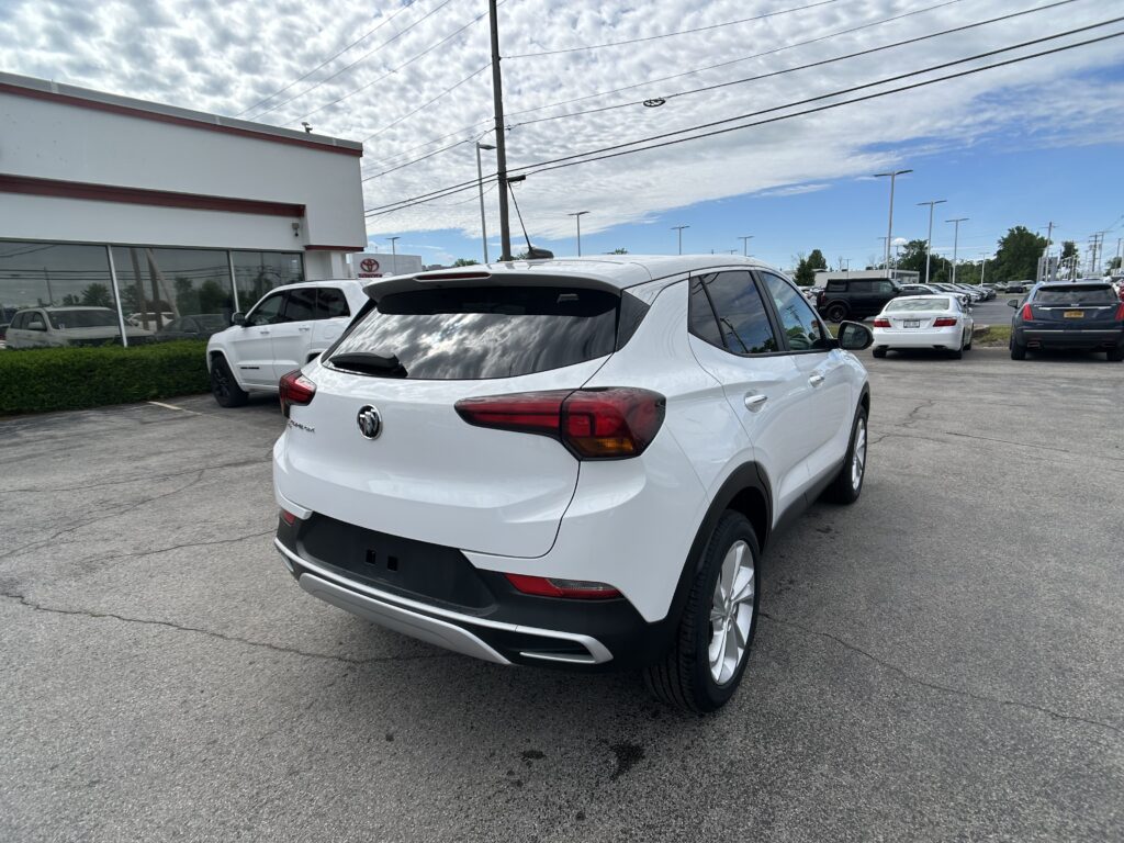 White Buick SUV in a car dealership lot with other cars and a white storefront on the left under a partly cloudy sky