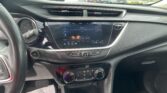 Car interior with a large touchscreen display, steering wheel, and climate controls; outside is a busy parking lot of cars. The windshield shows several vehicles.