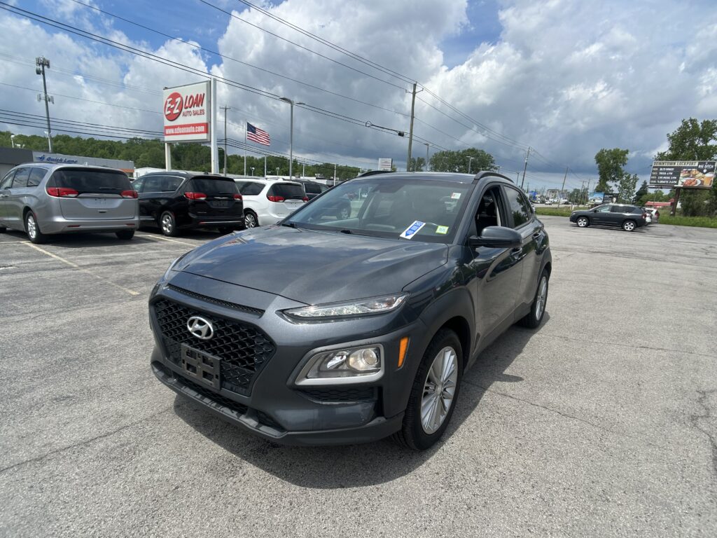 Gray Hyundai SUV in the foreground at a busy auto dealership lot with an EZ LOAN sign and American flag in the background