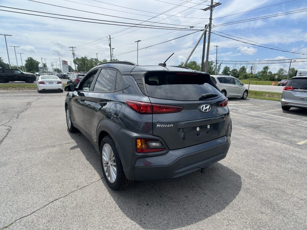 Rear-right view of a gray Hyundai Kona in a dealership parking lot, with other cars, power poles, and overhead cables visible.