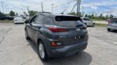 Rear-right view of a gray Hyundai Kona in a dealership parking lot, with other cars, power poles, and overhead cables visible.