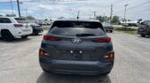 Rear view of a gray Hyundai Kona in a parking lot, other cars visible on both sides and power lines overhead under a partly cloudy sky.