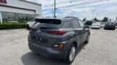 Rear view of a gray SUV in a car dealership lot, with a white storefront and many parked cars in the background, along power lines overhead.