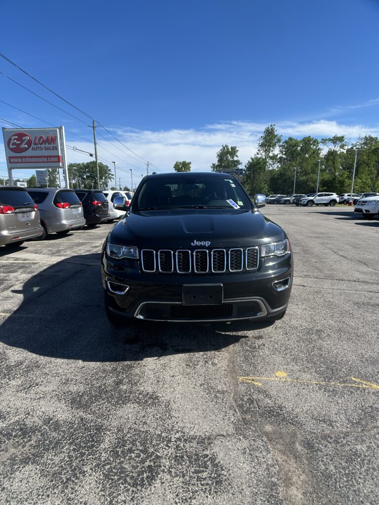 Front view of a dark Jeep SUV in a car dealership lot with rows of vehicles and an EZ Loan Auto Sales sign in the background
