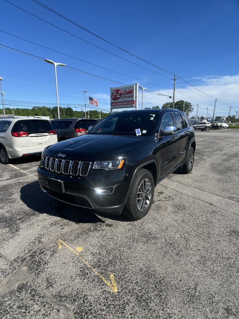 Black Jeep SUV in the dealership lot with an EZ Loan Auto Sales sign and an American flag in the background, under a clear blue sky.
