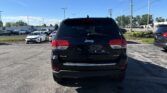 Rear view of a black Jeep SUV in a dealership parking lot under a clear blue sky with other cars around it.