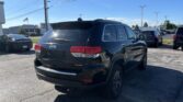 Black Jeep Grand Cherokee Limited parked in a dealership lot with other cars and power lines overhead under a clear blue sky.