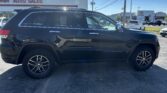 Side view of a black Jeep Grand Cherokee SUV parked in a car dealership lot, with a white auto sales building and a Toyota dealership in the background.