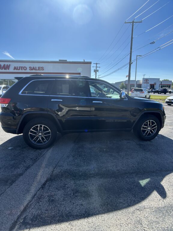 Side view of a black Jeep Grand Cherokee SUV parked in a car dealership lot, with a white auto sales building and a Toyota dealership in the background.