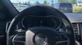 Driver’s-seat view inside a Jeep, steering wheel centered, instrument cluster visible, with a bright blue sky and other cars seen through the windshield in a parking lot.