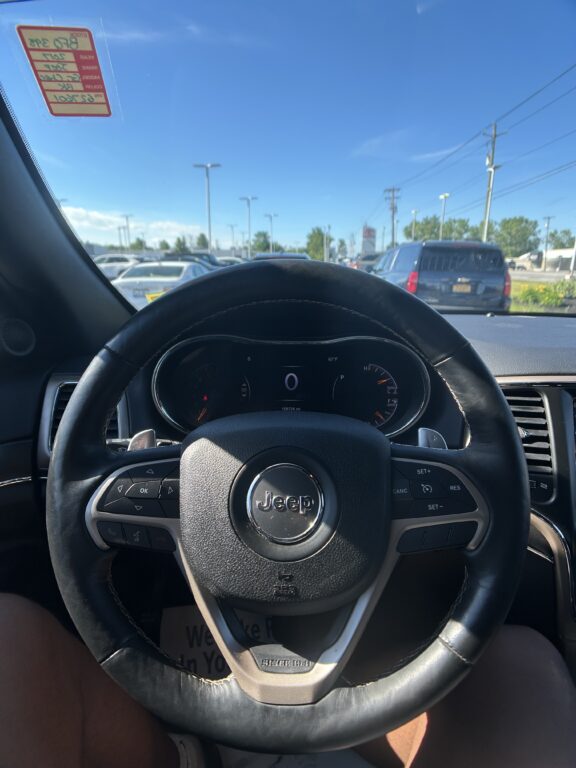 Driver’s-seat view inside a Jeep, steering wheel centered, instrument cluster visible, with a bright blue sky and other cars seen through the windshield in a parking lot.