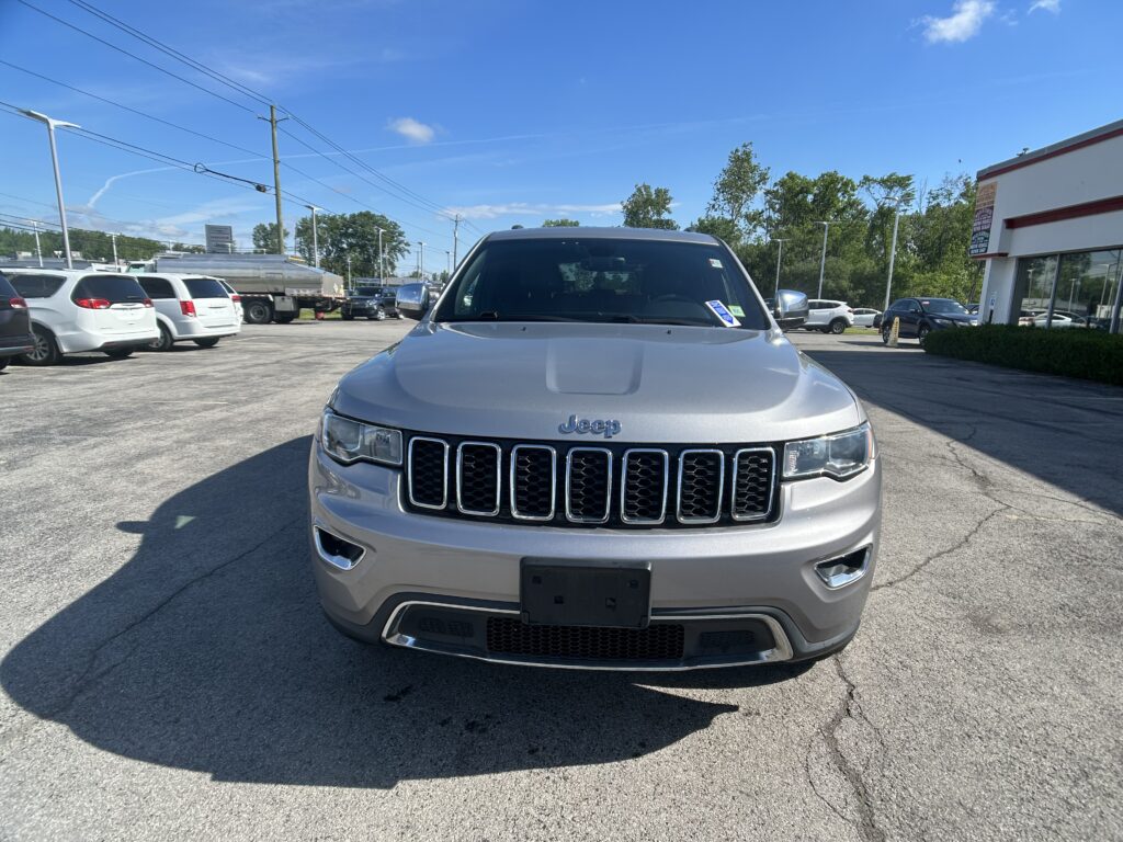 Front view of a silver Jeep SUV parked in a dealership lot with other cars and a building on the right under a clear blue sky, power lines above.
