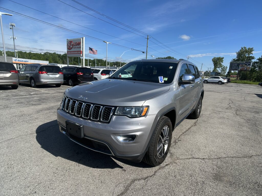 Silver Jeep SUV in a car lot with other vehicles, a large E-Z Loan Auto Sales sign, and an American flag against a bright blue sky.
