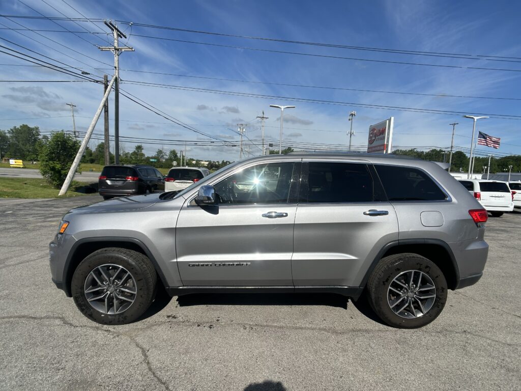 Silver Jeep Grand Cherokee parked in a parking lot with utility poles and power lines against a blue sky; American flag in the distance.