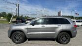 Silver Jeep Grand Cherokee parked in a parking lot with utility poles and power lines against a blue sky; American flag in the distance.