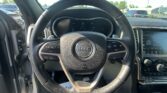 Driver's view of a car interior focused on the steering wheel and dashboard in a Jeep, with the infotainment screen to the right and a parking lot visible through the windshield in daylight.