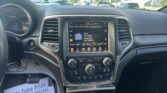 Interior of a car focusing on the center console: touchscreen radio screen showing music options, air vents, and climate knobs; view of a parking lot through the windshield; a flyer on the passenger side reads 'We Take Pride In Your Vehicle'.