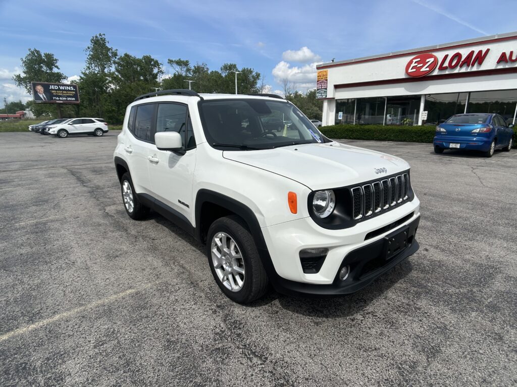 White Jeep SUV parked in a dealership lot with a blue building and other cars in the background under a partly cloudy sky.