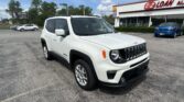 White Jeep SUV parked in a dealership lot with a blue building and other cars in the background under a partly cloudy sky.