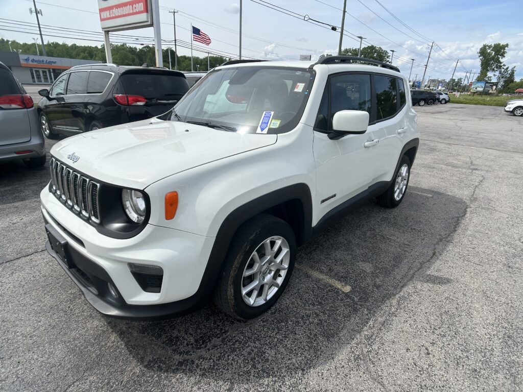 White Jeep Renegade in a busy car lot, with other vehicles and a dealership sign in the background and an American flag above.