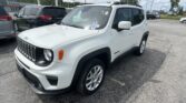 White Jeep Renegade in a busy car lot, with other vehicles and a dealership sign in the background and an American flag above.