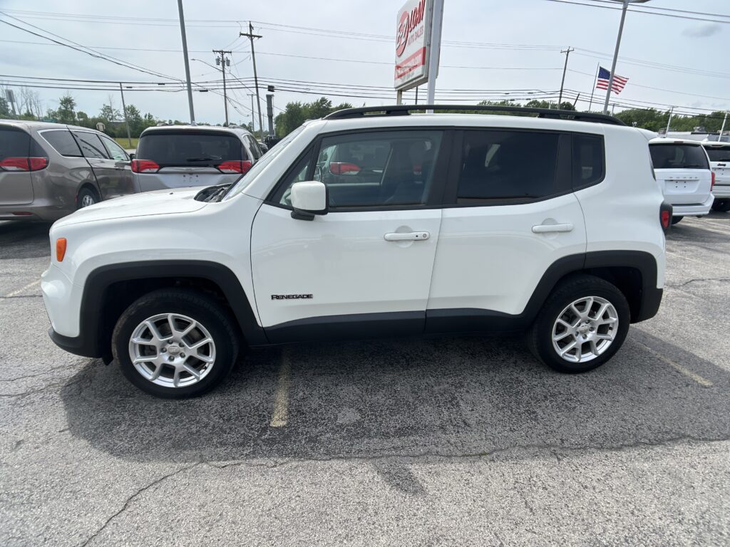 White Jeep Renegade parked in a car dealership lot, viewed from the passenger side with other vehicles nearby and an American flag in the background