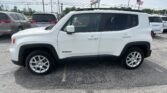 White Jeep Renegade parked in a car dealership lot, viewed from the passenger side with other vehicles nearby and an American flag in the background