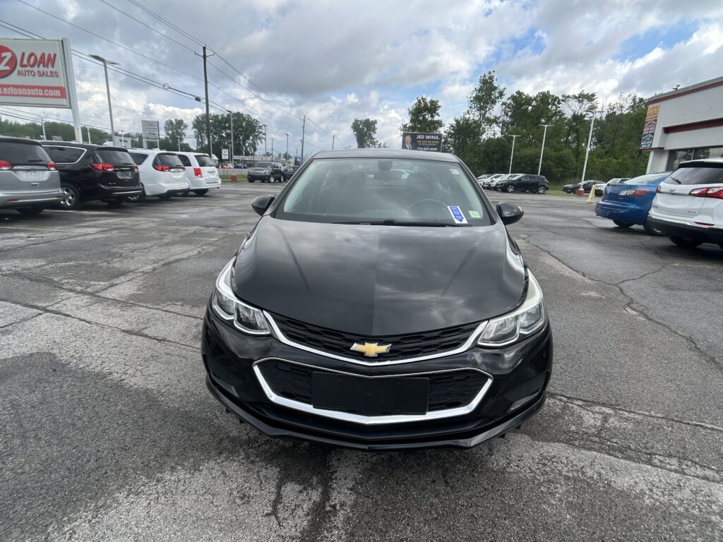 Front view of a black Chevrolet sedan in a crowded car dealership lot with other vehicles and signs nearby.