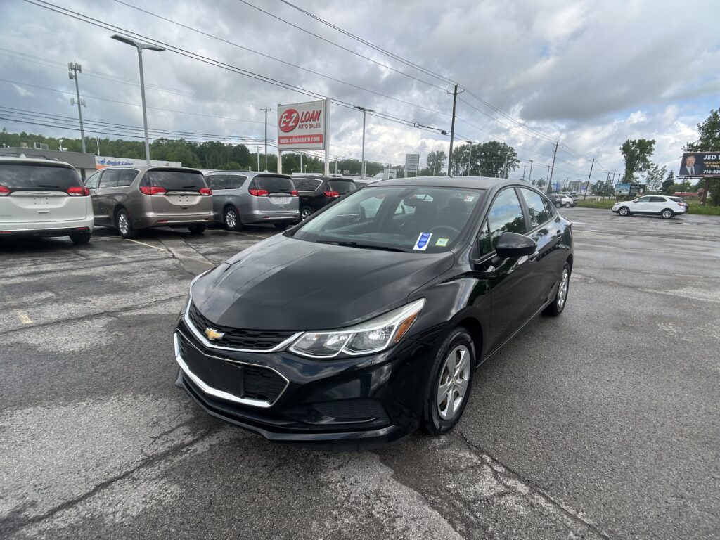 Black sedan in the foreground at a car dealership lot with rows of minivans and an E-Z Loan Auto Sales sign under a cloudy sky.