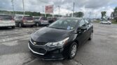 Black sedan in the foreground at a car dealership lot with rows of minivans and an E-Z Loan Auto Sales sign under a cloudy sky.