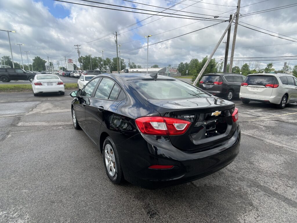 Black Chevrolet Cruze in the foreground of a dealership parking lot with other cars and tall power lines overhead under a cloudy sky.