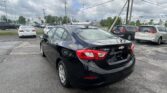 Black Chevrolet Cruze in the foreground of a dealership parking lot with other cars and tall power lines overhead under a cloudy sky.