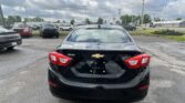 Rear view of a black Chevrolet Cruze in a car dealership or parking lot, with other vehicles and a cloudy sky above.