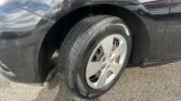 Close-up of a black car's front left wheel and fender, showing the tire and rim with reflections on the body surface.