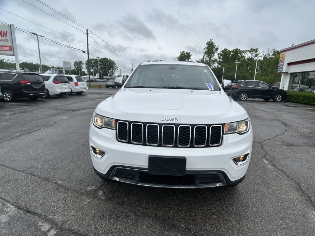 Front view of a white Jeep SUV parked in a car dealership lot, other cars and a building visible in the background under a cloudy sky.