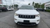 Front view of a white Jeep SUV parked in a car dealership lot, other cars and a building visible in the background under a cloudy sky.