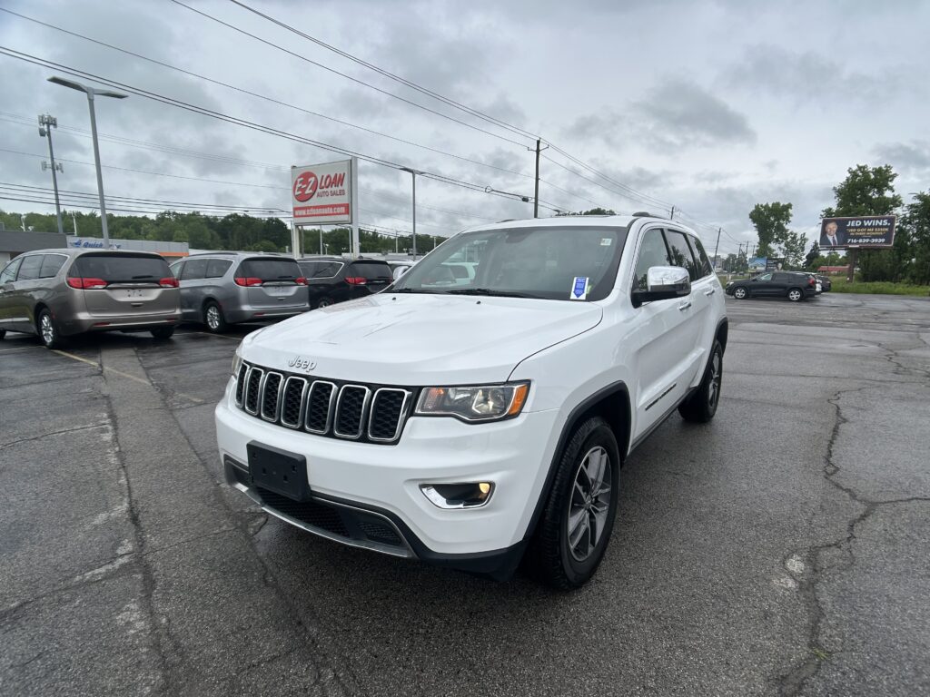 White Jeep SUV in a car dealership lot with other minivans and an Ez Loan sign in the background on a cloudy day.
