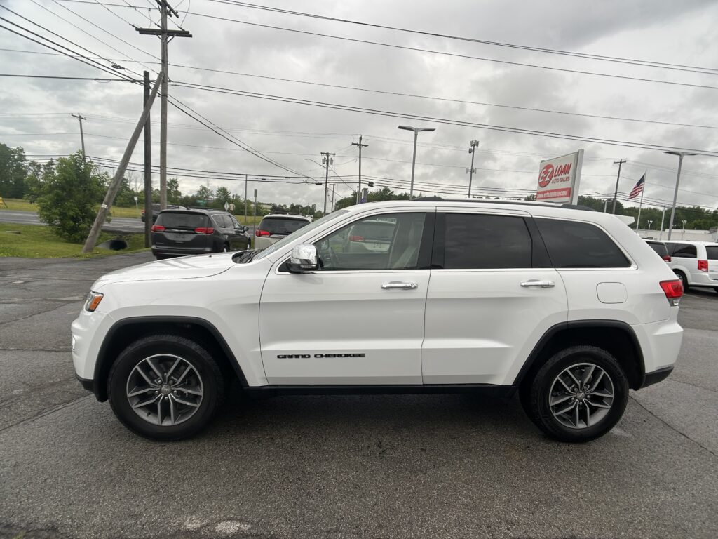 White Jeep Grand Cherokee SUV parked in a lot with utility poles, power lines, and a red EZ LOAN Auto Sales sign in the background
