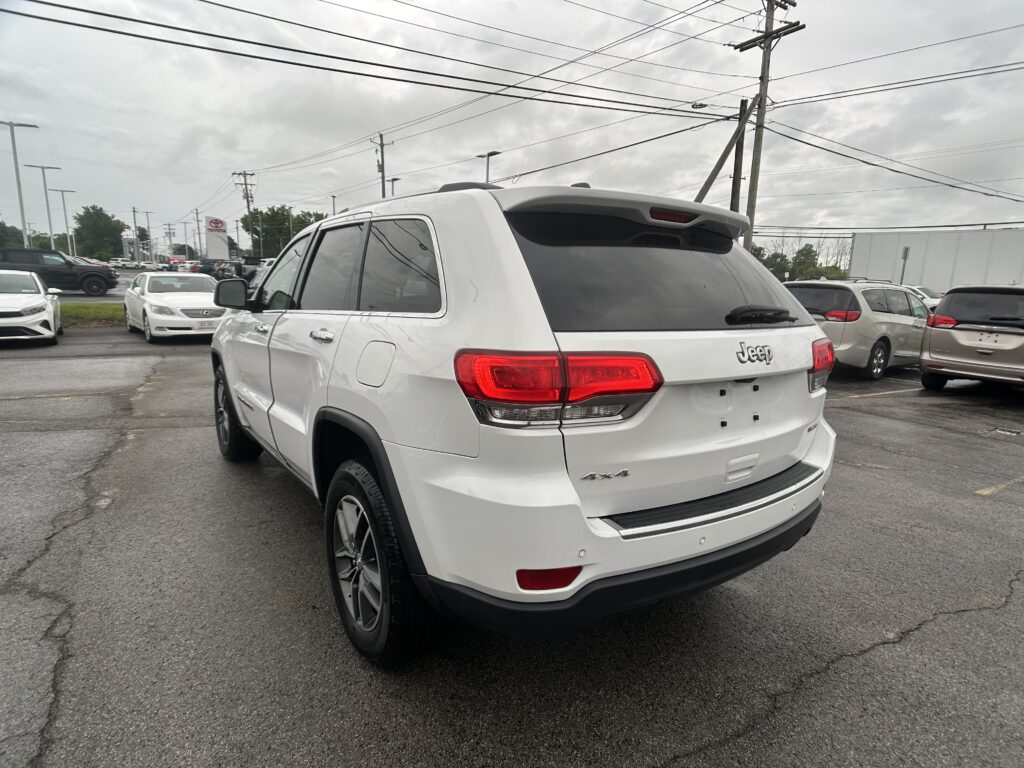 Rear three-quarter view of a white Jeep SUV in a car dealership lot with other cars and power lines in the background, cloudy sky above.