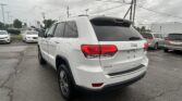 Rear three-quarter view of a white Jeep SUV in a car dealership lot with other cars and power lines in the background, cloudy sky above.