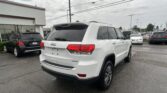 Rear three-quarters view of a white Jeep Grand Cherokee Limited in a dealership lot, with other cars and a white building to the left under a cloudy sky.