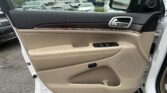 Interior of a beige car door with black upper panel, wood trim, chrome handle, and window controls; view of a rainy dealership lot outside the window.