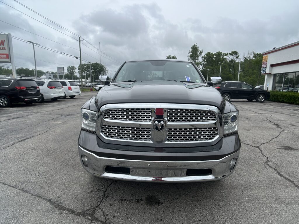 Front view of a silver RAM pickup truck in a dealership lot, with other cars and a building in the background under a cloudy sky.