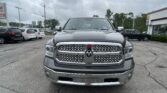 Front view of a silver RAM pickup truck in a dealership lot, with other cars and a building in the background under a cloudy sky.