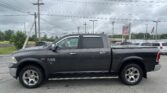 Gray RAM 1500 pickup in a dealership parking lot with a cloudy sky and power lines in the background, viewed from the passenger side.